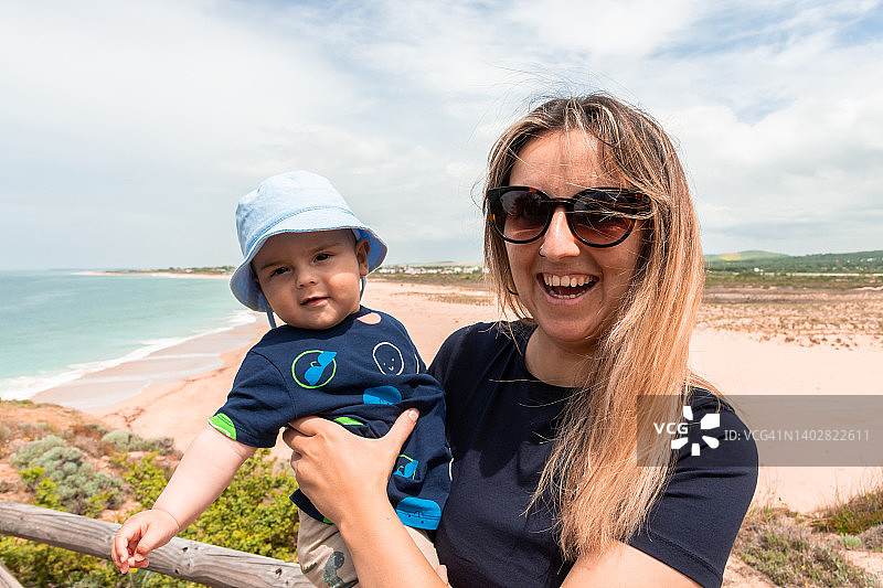 海滩背景下抱着宝宝的快乐母亲的夏日肖像图片素材