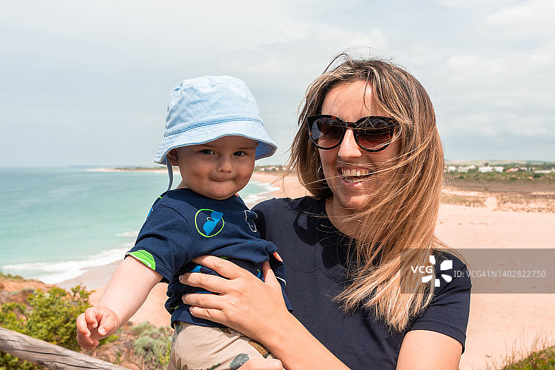 海滩背景下抱着宝宝的快乐母亲的夏日肖像图片素材