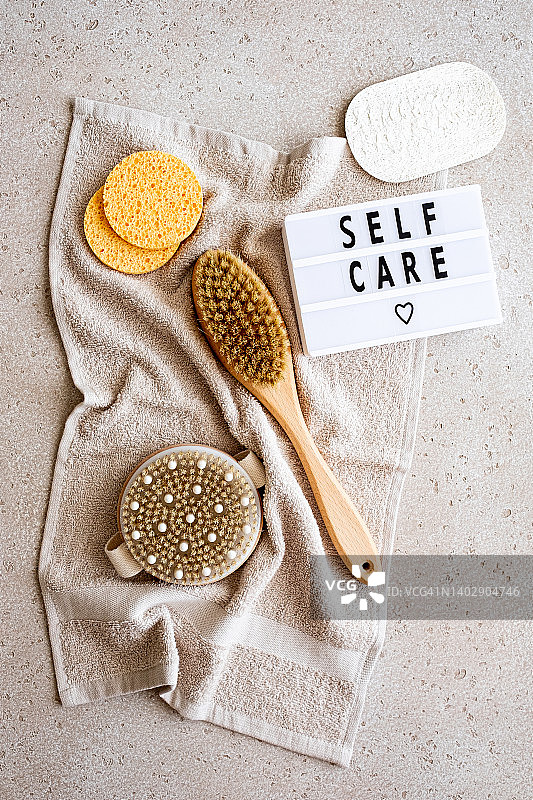 家庭水疗设备（棉毛巾、木制身体刷、清洁海绵、丝瓜络）和带有“呵护自己”字样的灯箱，混凝土背景，俯视。可持续生活方式。零浪费概念图片素材