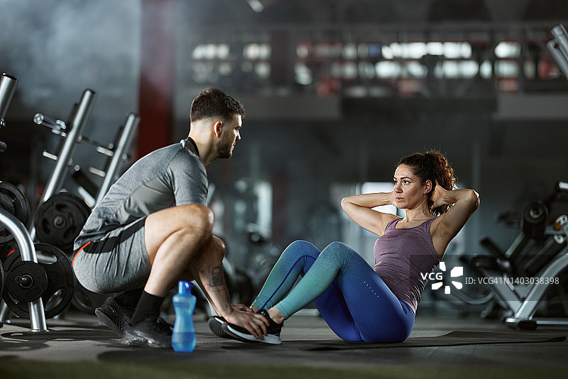 年轻男教练在健身房协助运动型女人做仰卧起坐图片素材