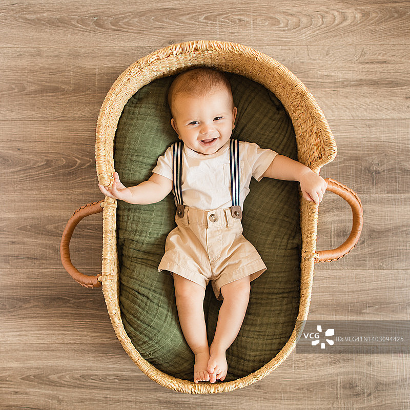 一位25周大的男婴穿着白色衬衫、海军条纹背带和卡其色短裤，躺在舒适的绿色棉毯中，在一个海草摩西篮子里，周围环绕着热带室内植物，营造出波西米亚风情的热带氛围图片素材