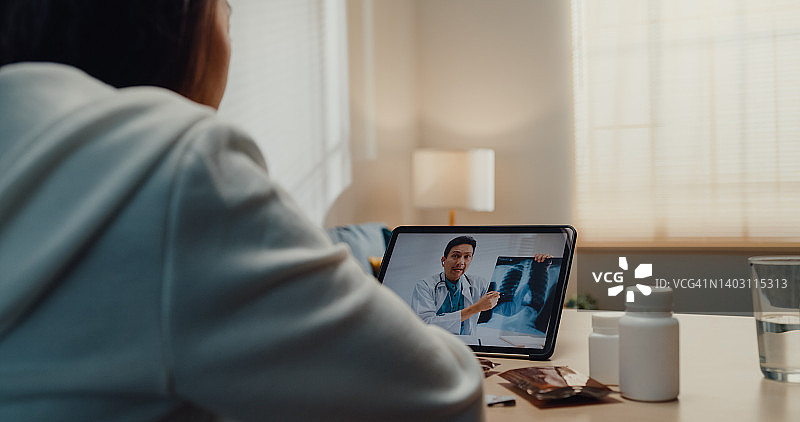 生病的年轻亚洲女人在家中客厅使用平板电脑与医生交谈图片素材