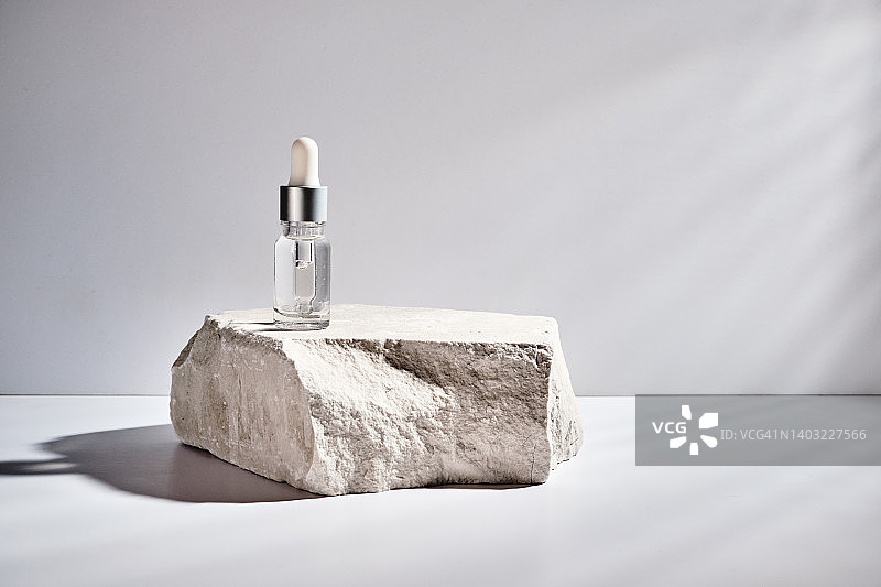 带有透明化妆品凝胶精华质地的化妆品瓶，立于带有硬阴影的岩石基座上。用于滋润和美白皮肤的化妆品美容概念图片素材