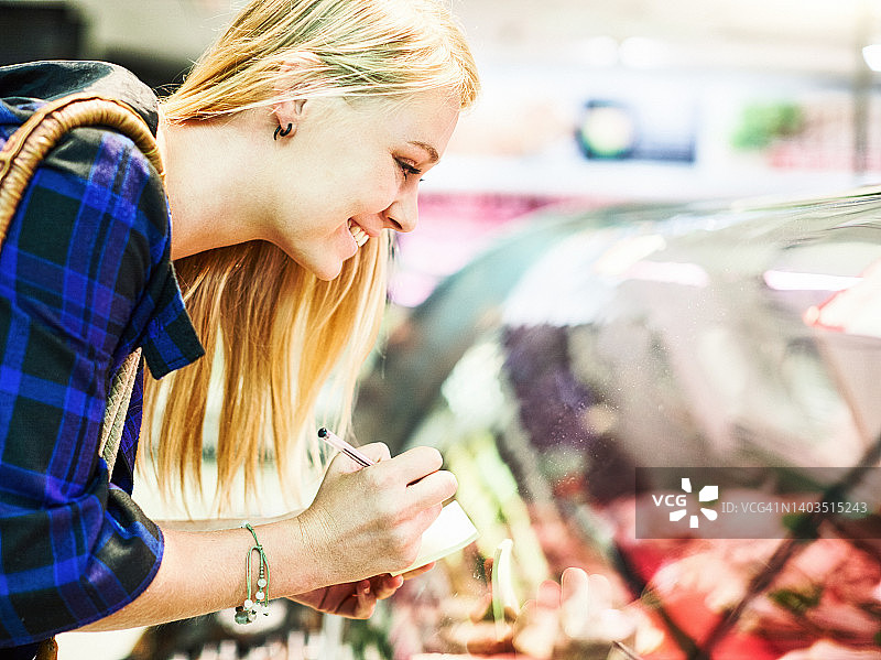 年轻金发女子在超市购物，在熟食区挑选玻璃柜下的商品，微笑并查看购物清单图片素材