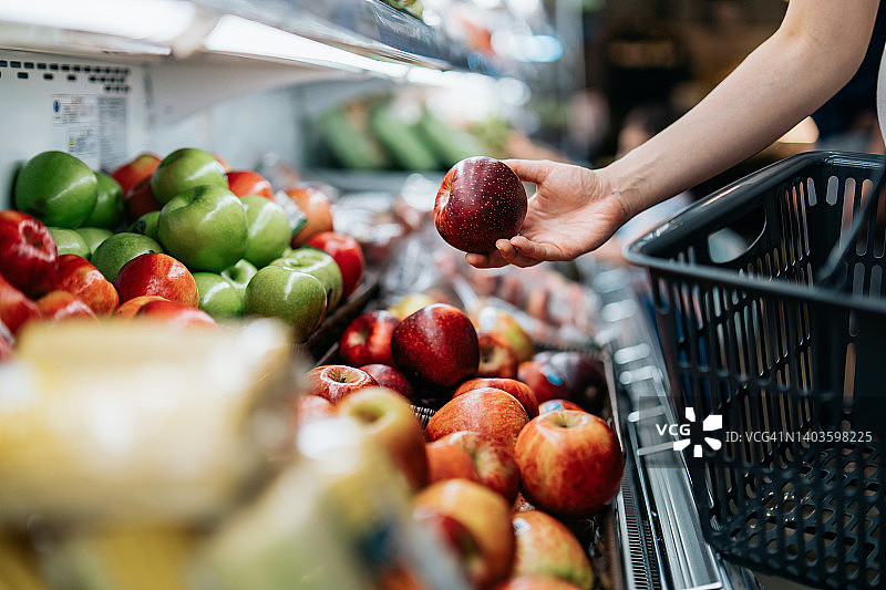 年轻亚裔女性在超市挑选新鲜有机水果的局部照片。她在农产品区挑选一个红苹果。日常食品采购，健康生活和饮食方式图片素材