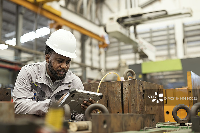 制造业中注塑模具资产的集中管理。非裔美国模具工程师在模具存储区使用数字平板电脑交叉检查模具资产，并使用管理工具软件进行比较。图片素材