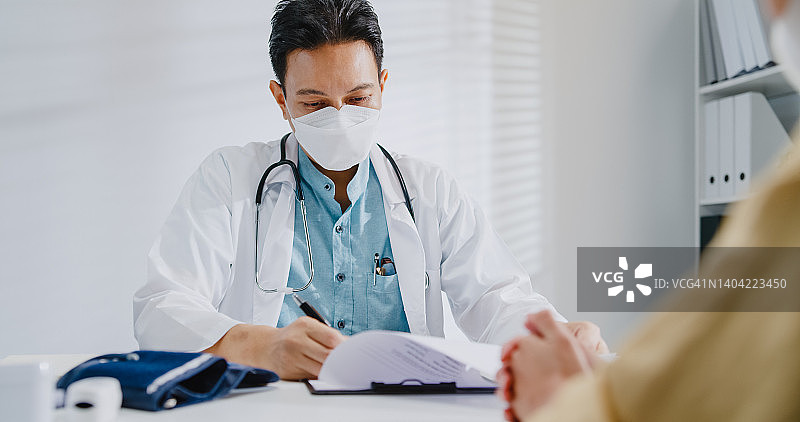 年长的亚洲男医生在诊所与女病人咨询时查看医疗结果，佩戴防护口罩图片素材
