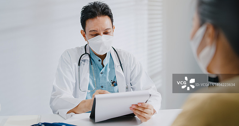 年长的亚洲男医生在诊所与女病人咨询时查看医疗结果，佩戴防护口罩图片素材