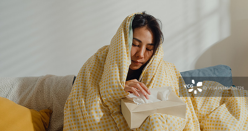 年轻亚洲女性感冒，在客厅沙发上用纸巾擦鼻子、打喷嚏图片素材