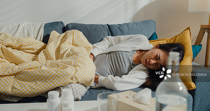 生病的年轻亚洲女人腹部剧烈疼痛，躺在家里客厅沙发上的毯子下图片素材