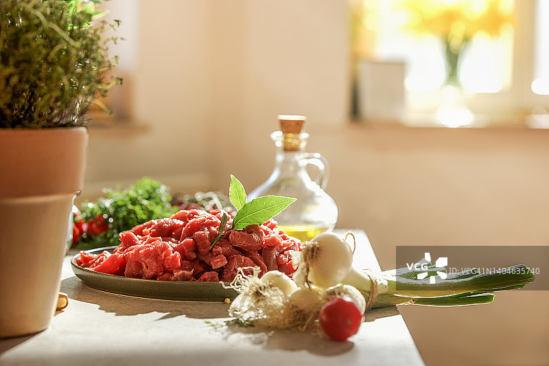 现代厨房静物，桌子上放着切好的生肉和烹饪原料：葱、番茄、盆栽香草和橄榄油，窗户内部背景图片素材