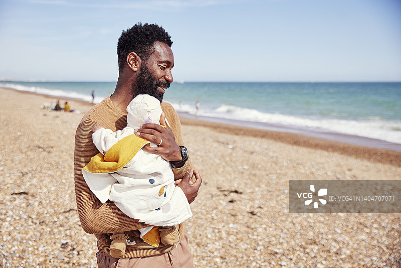 父亲在海滩上抱着他的宝宝，紧贴胸前，淡季家庭公路旅行图片素材