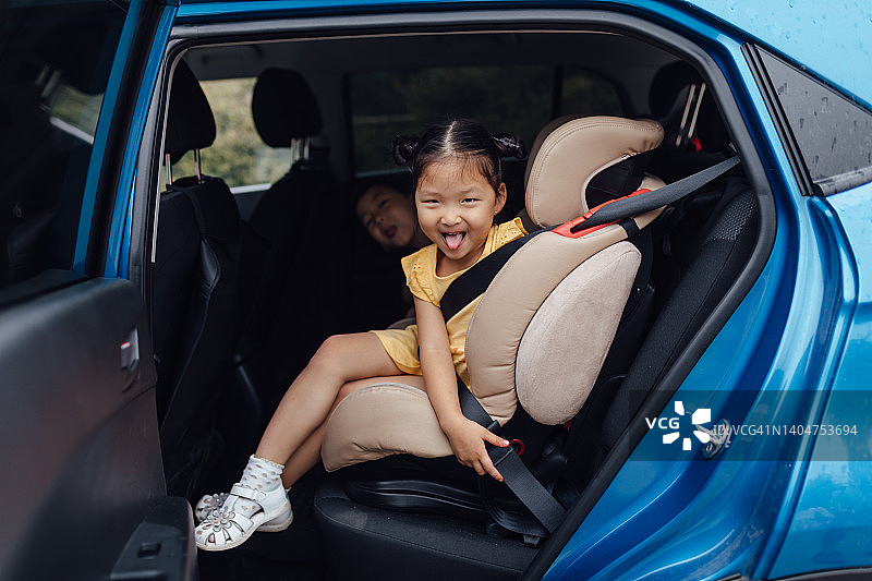 穿着黄色连衣裙的亚洲小女孩坐在儿童汽车座椅上，和哥哥一起看向镜头，吐着舌头微笑图片素材