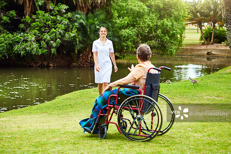 微笑的护士走向坐在轮椅上的老妇人，她身旁是倒映着茂盛树叶的绿色草坪和池塘图片素材