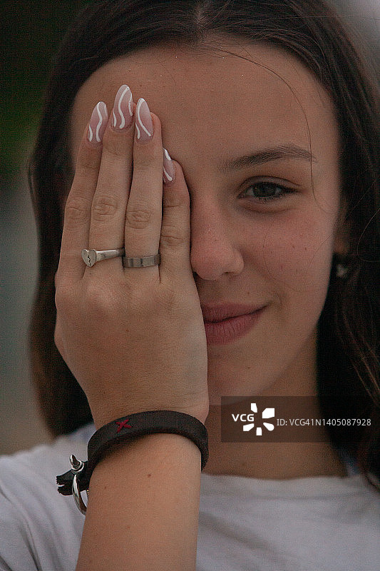 身穿白色T恤的年轻女子，长长的棕色头发披散着，用一只手遮住一只眼睛，展示着非常长的华丽指甲和戒指和手镯形式的珠宝图片素材