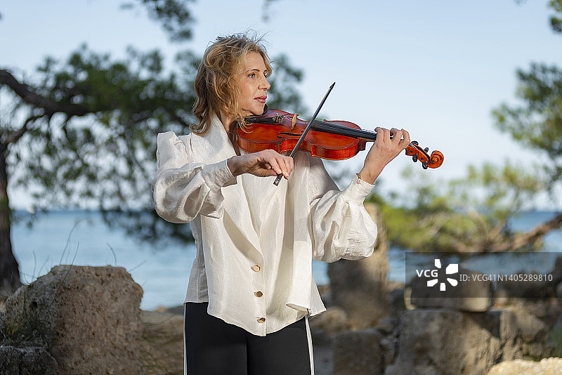 在海边，在树林里，在夏天，一位穿着白色衬衫的金发中年妇女在演奏小提琴图片素材