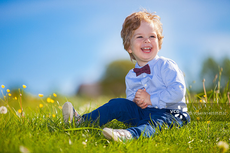系着领结坐在绿色草地上的可爱微笑男婴。有趣的快乐男孩特写肖像。恶作剧的小孩在绿草地上玩耍并看向一侧图片素材