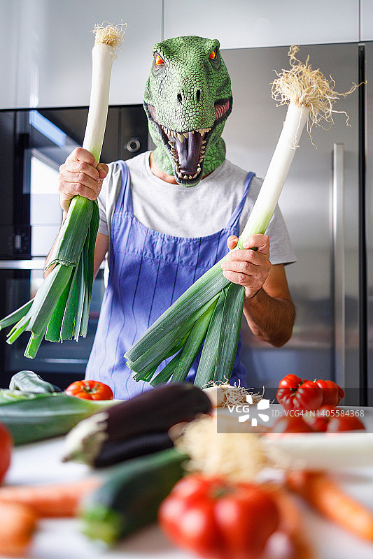 戴着恐龙面具的男人在厨房里拿着韭菜，桌子上摆满了蔬菜，健康饮食概念图片素材