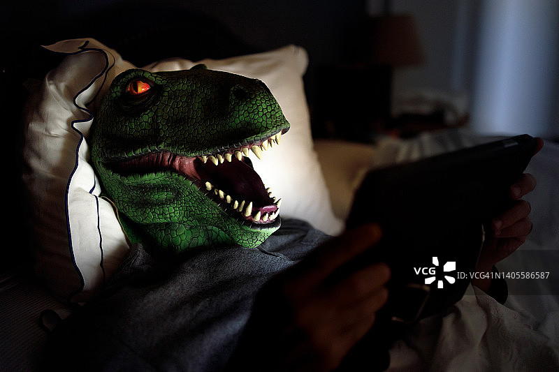 戴着恐龙面具的男人躺在床上，用数字平板电脑阅读图片素材