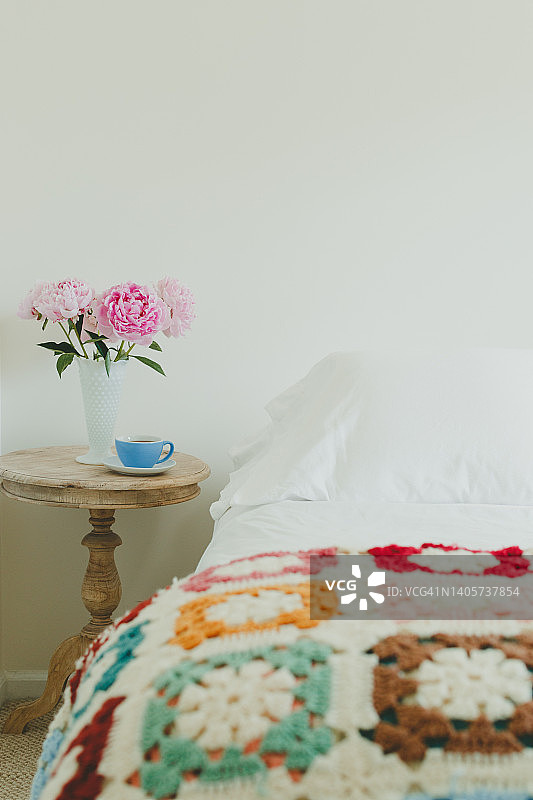 带有奶奶方块彩色阿富汗毯的床，以及带有粉色牡丹花瓶和蓝色茶杯的边桌图片素材