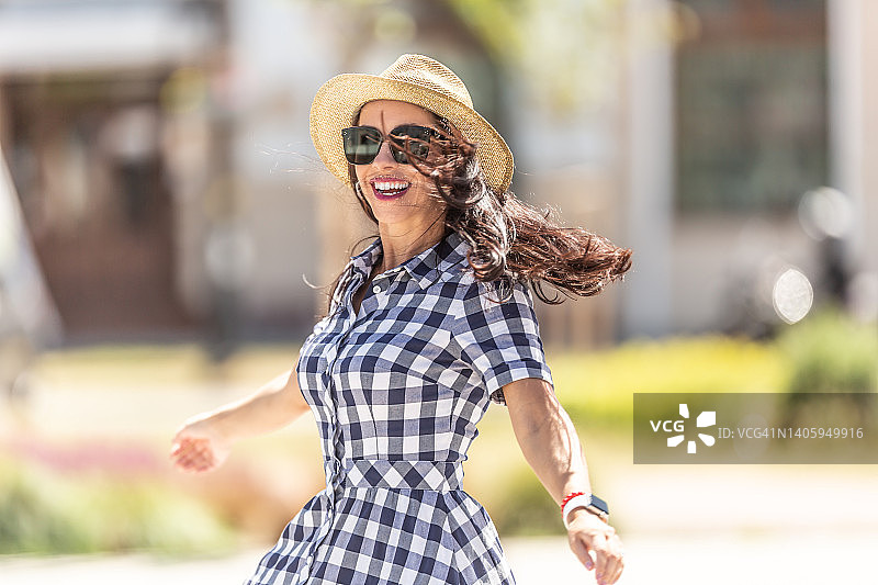 美丽的黑发女人穿着时尚的连衣裙、眼镜和帽子，享受阳光明媚的夏日图片素材