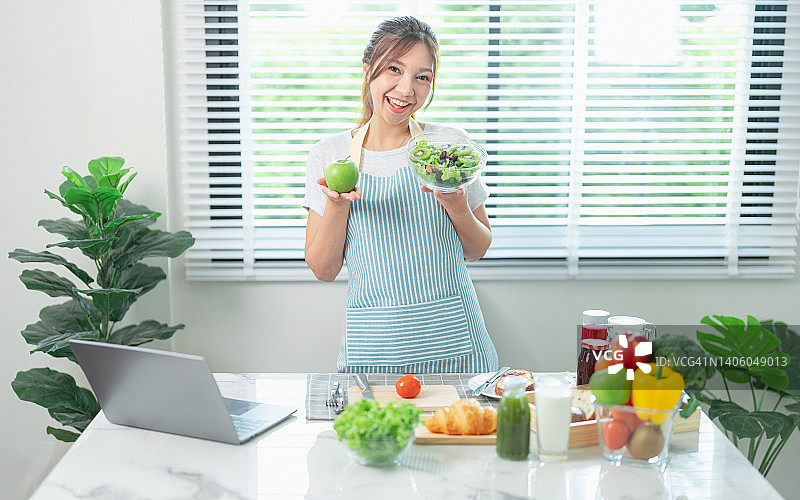 美丽的亚洲博主正在吃自制新鲜沙拉早餐，并录制在线频道直播视频。运动型美女在家中厨房一边吃健康沙拉，一边通过笔记本电脑进行在线视频通话图片素材