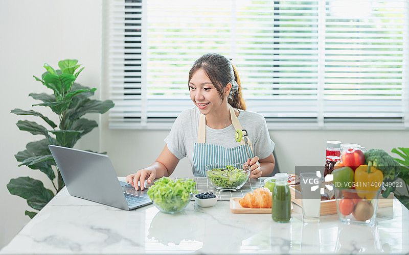 美丽的亚洲博主正在吃自制新鲜沙拉早餐，并录制在线频道直播视频。运动型美女在家中厨房一边吃健康沙拉，一边通过笔记本电脑进行在线视频通话图片素材