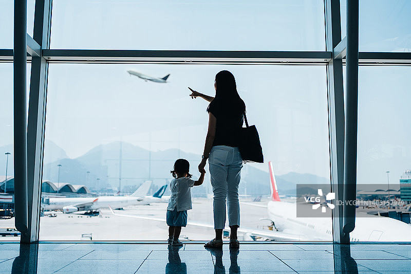 在机场等待起飞时，年轻的亚洲母亲牵着可爱的小女儿的手，隔着窗户看着飞机的背影。家庭旅行和度假概念图片素材