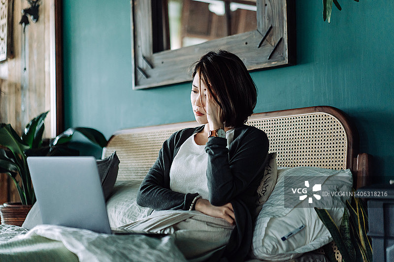 担心的年轻亚洲女人手扶着头，在家里的床上使用笔记本电脑，看起来很担心，有压力图片素材