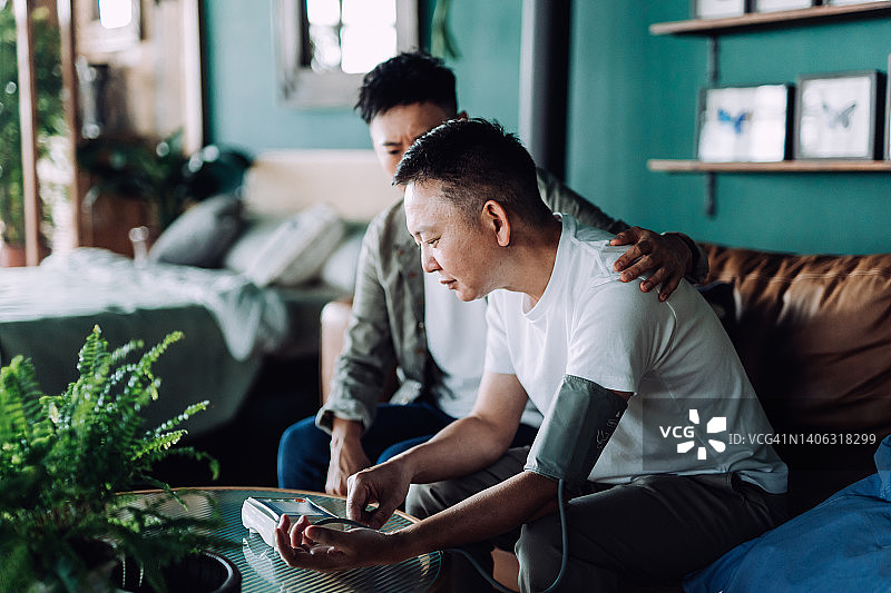年长的亚洲男人在家与儿子一起检查血压，看起来很担心，儿子关心他退休的父亲，手搭在他的肩膀上，安慰和慰问他的父亲，家庭爱，关怀，老年和医疗保健概念图片素材