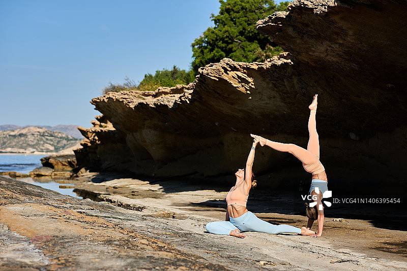 两位女性在岩石海滩上进行阿克罗瑜伽，展示头倒立和鸽子式瑜伽图片素材