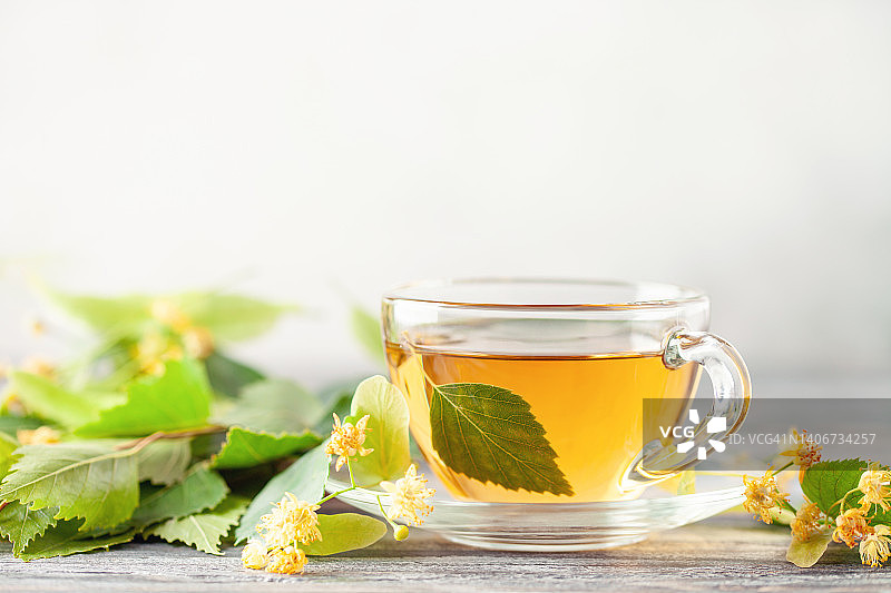 绿茶和菩提花以及桦树叶的玻璃茶杯在木制背景上。草药热饮图片素材