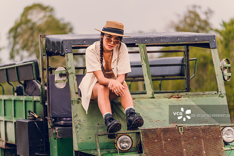 穿着Safari主题连衣裙、戴帽子、眼镜和珠宝的时尚女人站在田野和树木旁的生锈卡车旁图片素材
