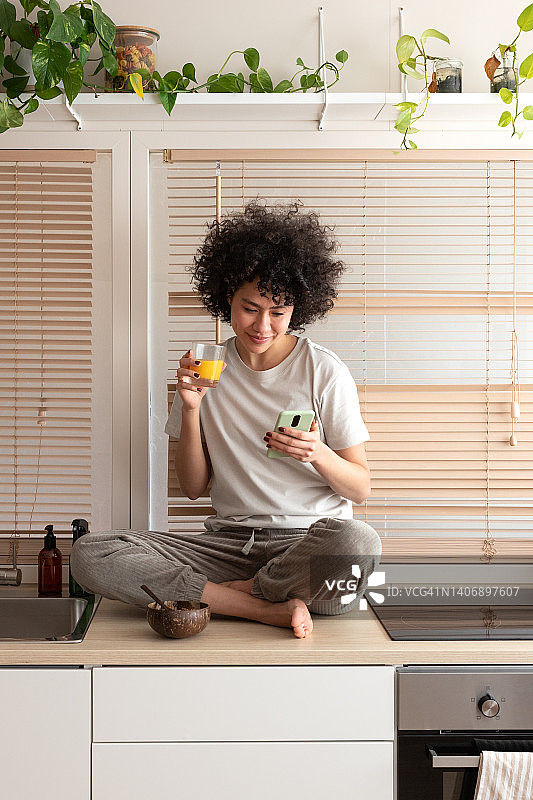 年轻的混血女人在厨房台面上看手机，吃着健康的早餐，喝着橙汁。竖版图片。图片素材