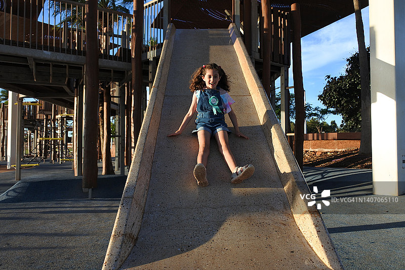 快乐的年轻澳大利亚女孩在操场上的滑梯上滑滑梯图片素材