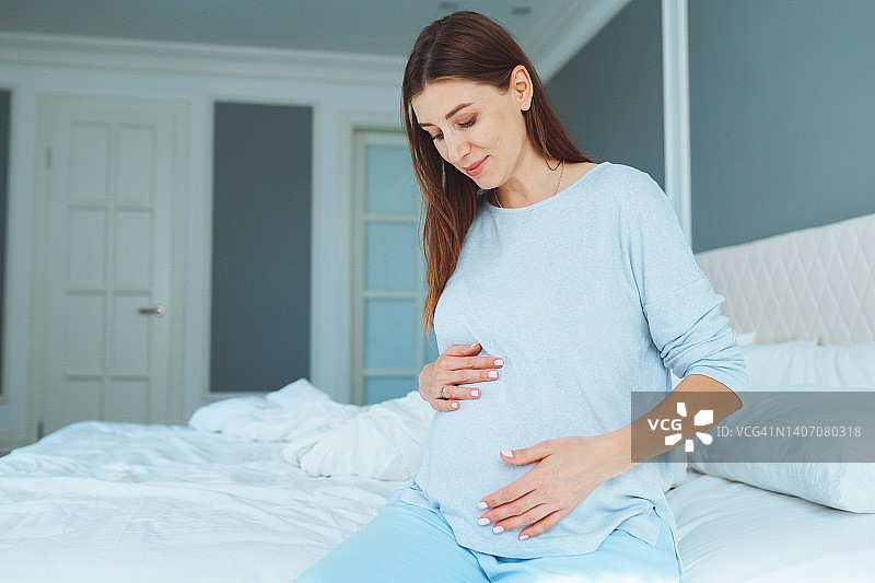 平静的孕妇轻轻抚摸着肚子，呵护着未出生的宝宝，坐在床上图片素材