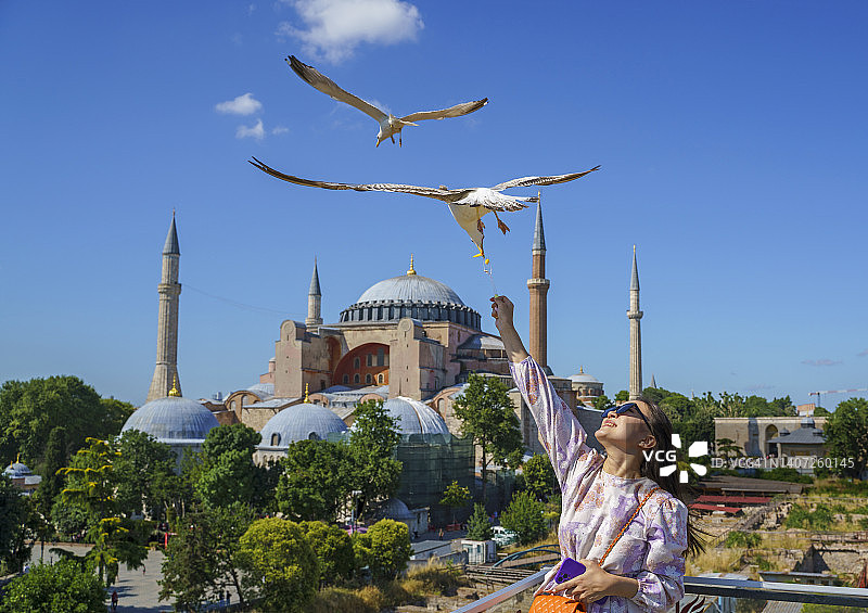 年轻的亚洲女人在屋顶喂海鸥，背景是土耳其伊斯坦布尔圣索菲亚大教堂图片素材