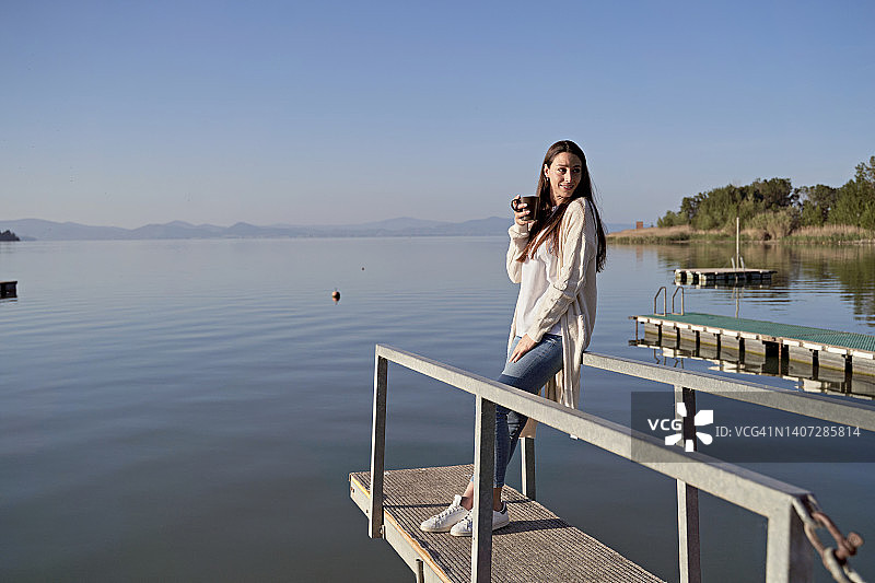 女人端着咖啡杯站在湖边的码头上图片素材