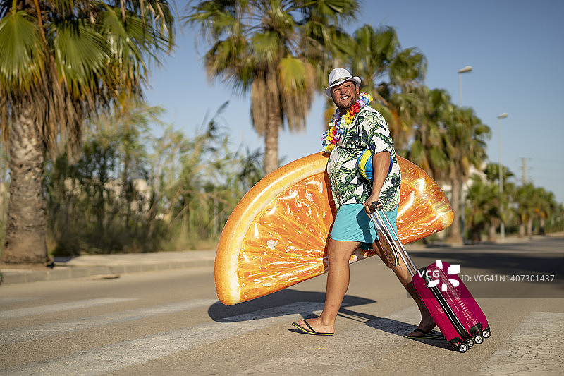 微笑的男人拿着橙色泳池漂浮物和行李箱过马路图片素材