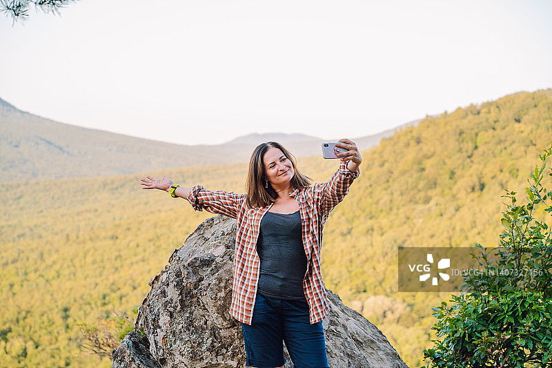 中年妇女在国外旅行，站在石崖附近，用智能手机对着山地水平线自拍图片素材
