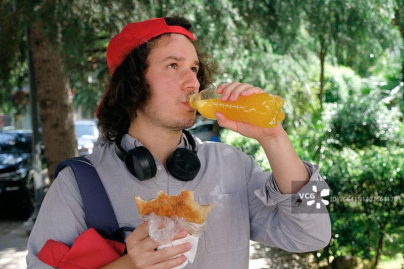 男人在户外手持格鲁吉亚哈恰普里面包，一边吃一边喝碳酸饮料，学生或游客吃外卖食品，青少年吃酥皮点心，快餐概念，在旅途中了解格鲁吉亚美食图片素材
