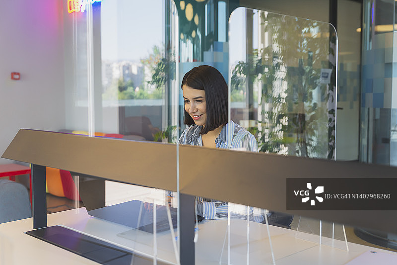 疫情期间，年轻女商人在现代办公室的有机玻璃防护墙后微笑工作图片素材