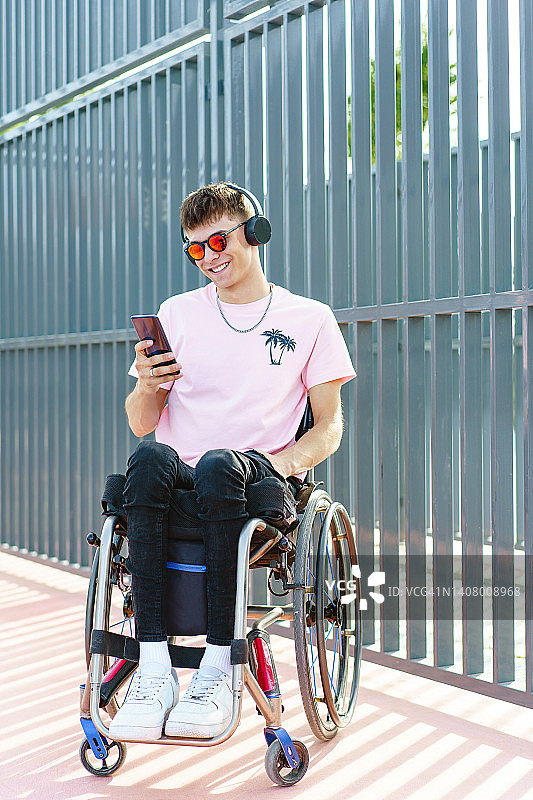 穿着都市粉色衣服坐在轮椅上，在户外使用智能手机的男人的全身水平视角图片素材