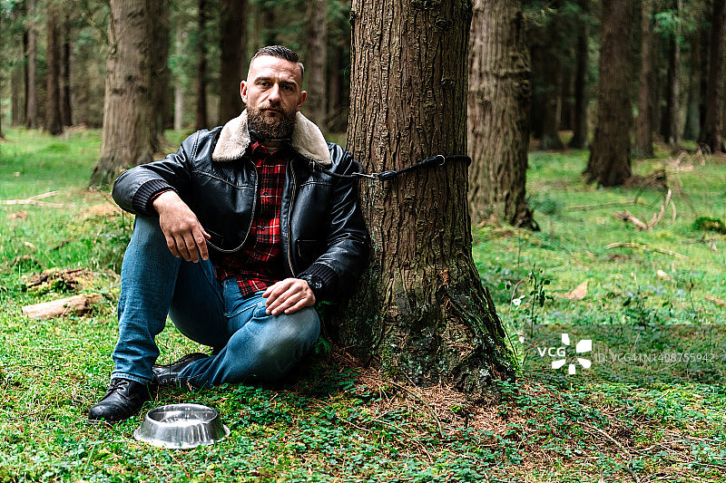 被绑在森林里树上的英俊男人，旁边放着一个空狗碗，谴责遗弃动物行为图片素材
