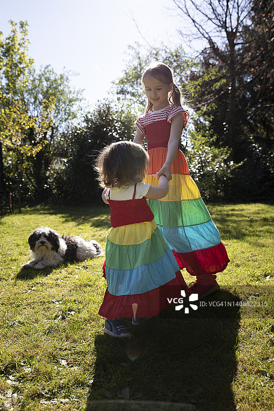 幼儿和姐姐穿着彩虹色连衣裙在花园草地上跳舞，宠物狗在背景中观看图片素材