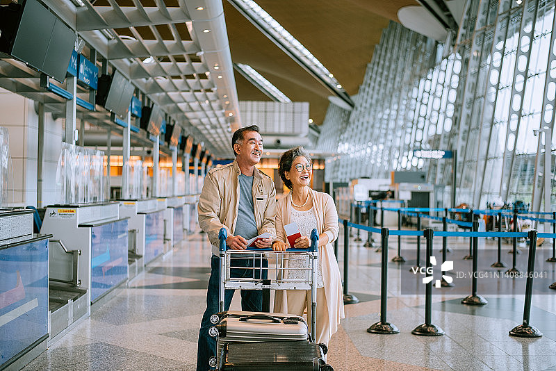 老年夫妇在机场航站楼拉着行李箱走过窗边，隔着玻璃窗观看飞机图片素材