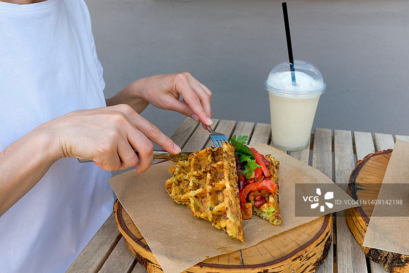 在户外咖啡馆或餐厅的木桌旁吃华夫饼和烤蔬菜的女人。早餐，商务午餐。素食和纯素食品的概念。旁边放着奶昔、柠檬水。图片素材