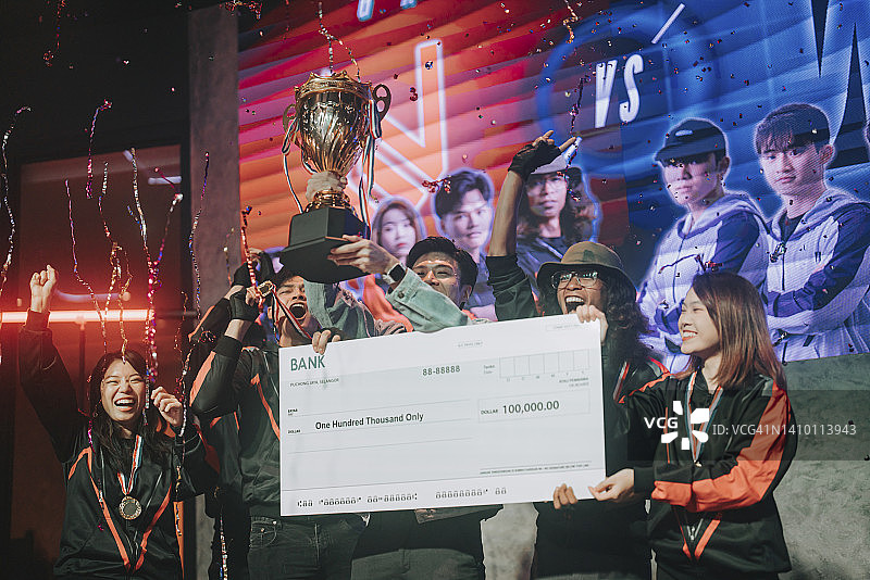亚洲电竞团队手持奖杯模型支票，在舞台上欢呼雀跃，庆祝赢得视频游戏总决赛。视频游戏锦标赛竞技场。电子游戏锦标赛赛事图片素材