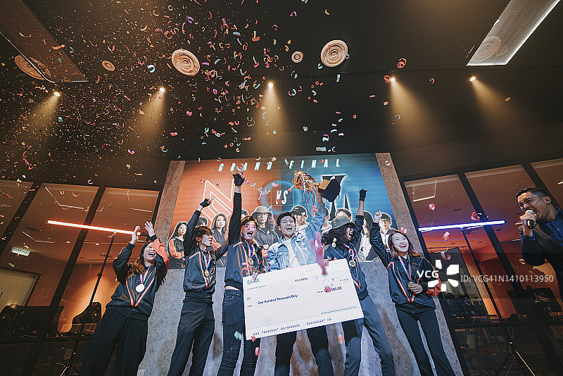 亚洲电竞团队手持奖杯模型支票，在舞台上欢呼雀跃，庆祝赢得视频游戏总决赛。视频游戏锦标赛竞技场。电子游戏锦标赛赛事图片素材