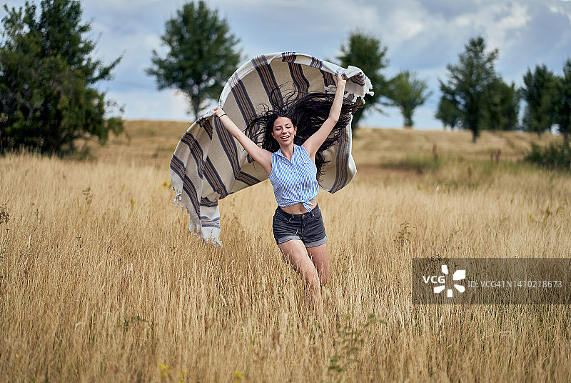 微笑的年轻女子在绿草地上奔跑，手臂在空中挥舞着一条长长的白色围巾。快乐、积极和健康的女士享受自由和幸福图片素材